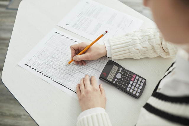 Student filling out a test answer sheet with a pencil, using a scientific calculator on a desk during an exam.